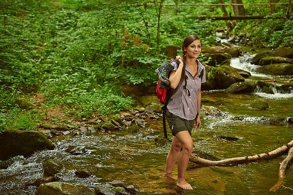 Frau mit Rucksack durchquert barfuß einen Bach im Wald