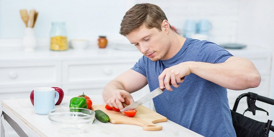 Mann im Rollstuhl sitzt in der Küche und schneidet Gemüse auf einem Brett