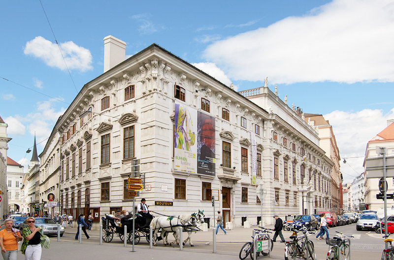 Zu sehen ist die Frontalansich auf das Theatermuseum in Wien, ein Gebäude aus der Jahrhundertwende davor der Lobkowitzplatz mit vielen Menschen und einer Kutsche.