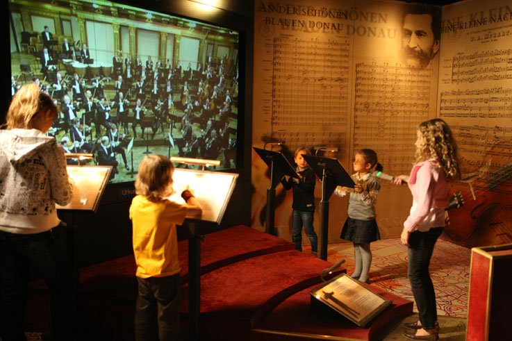 5 junge Kinder dirigieren vor einer Leinwand, auf der ein Konzert abgespielt wird