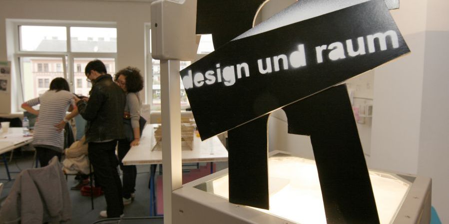 Werkstätte Design mit drei Menschen im Hintergrund und schwarzem Schild mit Inschrift design und raum davor