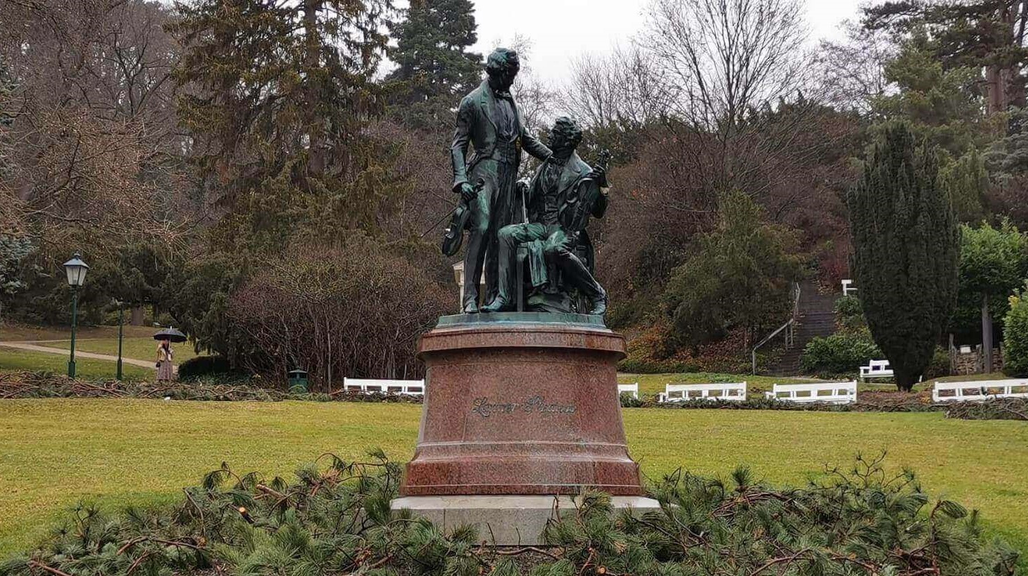 Zwei oxidierte Bronzestatuen von Joseph Lanner und Johann Strauß auf einem runden Fundament umringt von Ästen von Nadelbäumen, während im Hintergrund eine weite Wiese und Bäume zu sehen sind.