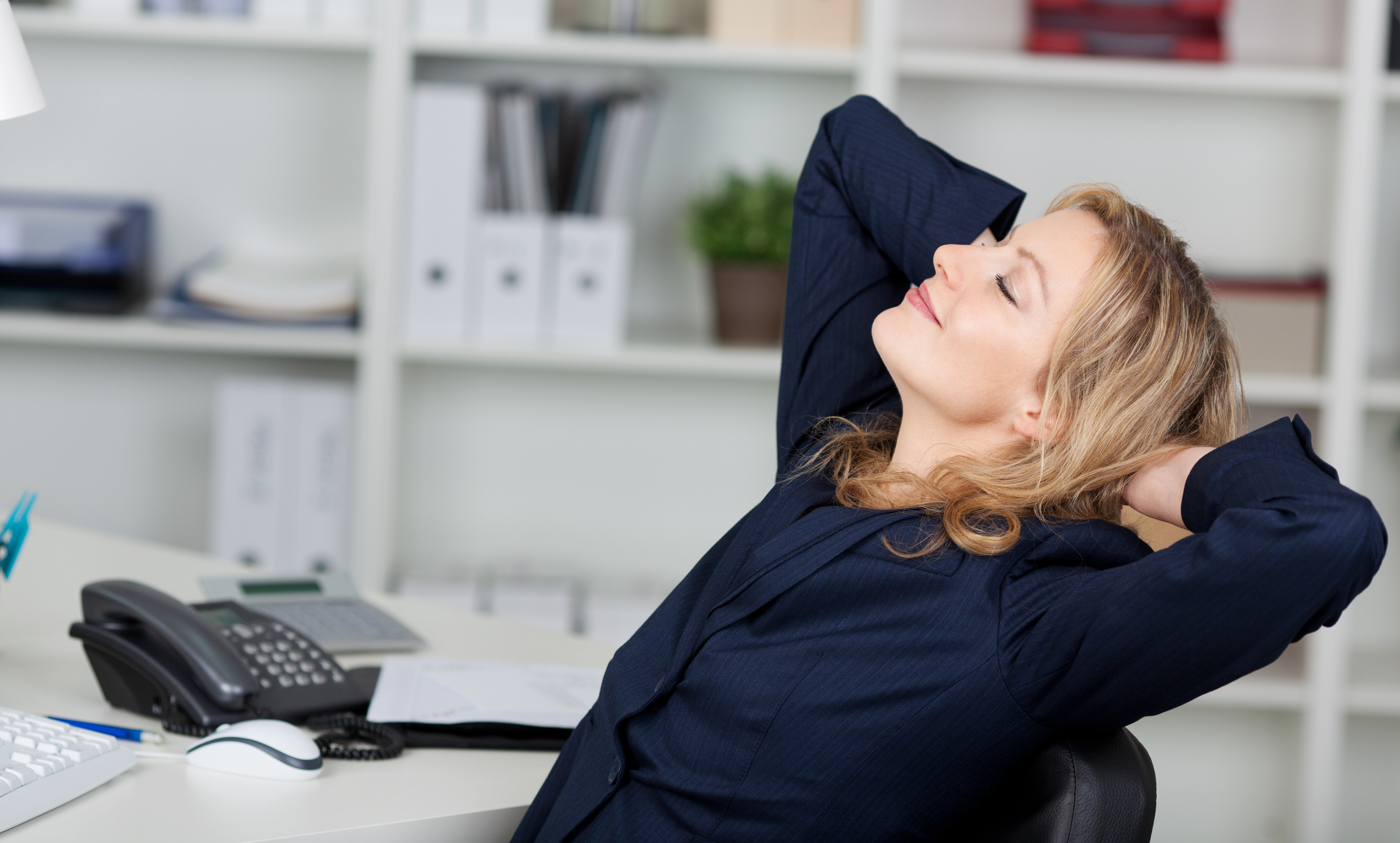 Frau die entspannt auf einem Bürosessel sitzt und die Arme hinter dem Kopf verschränkt hält
