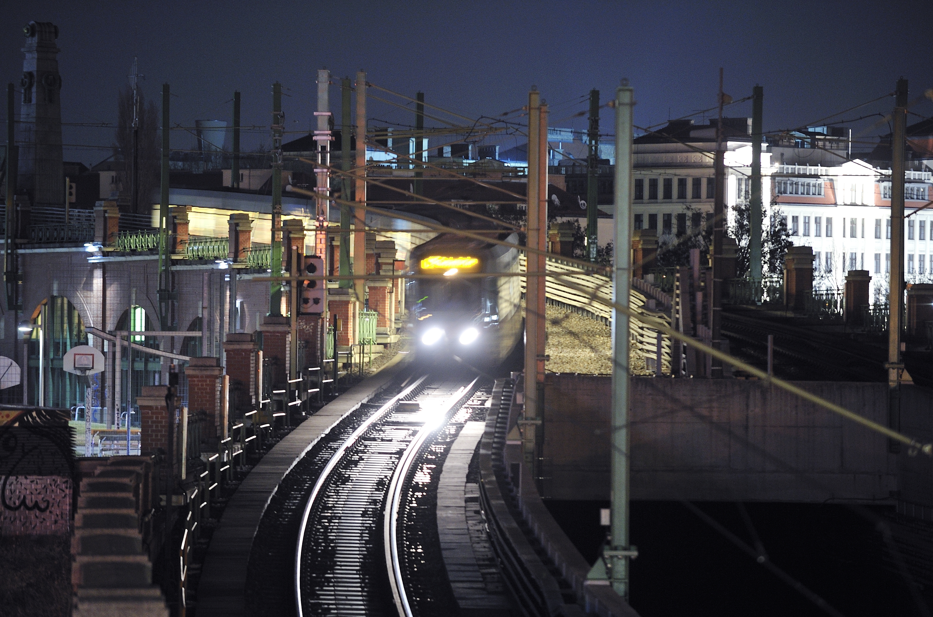 Bild der U-Bahn-Linie U6 bei Nachtbetrieb. Der Zug hat die Scheinwerfer an und ist in der Nähe der Station Längenfeldgasse unterwegs. Im Hintergrund befinden sich beleuchtete Häuser und die Gürtelbögen, sowie ein Basketballkorb und Graffitis.