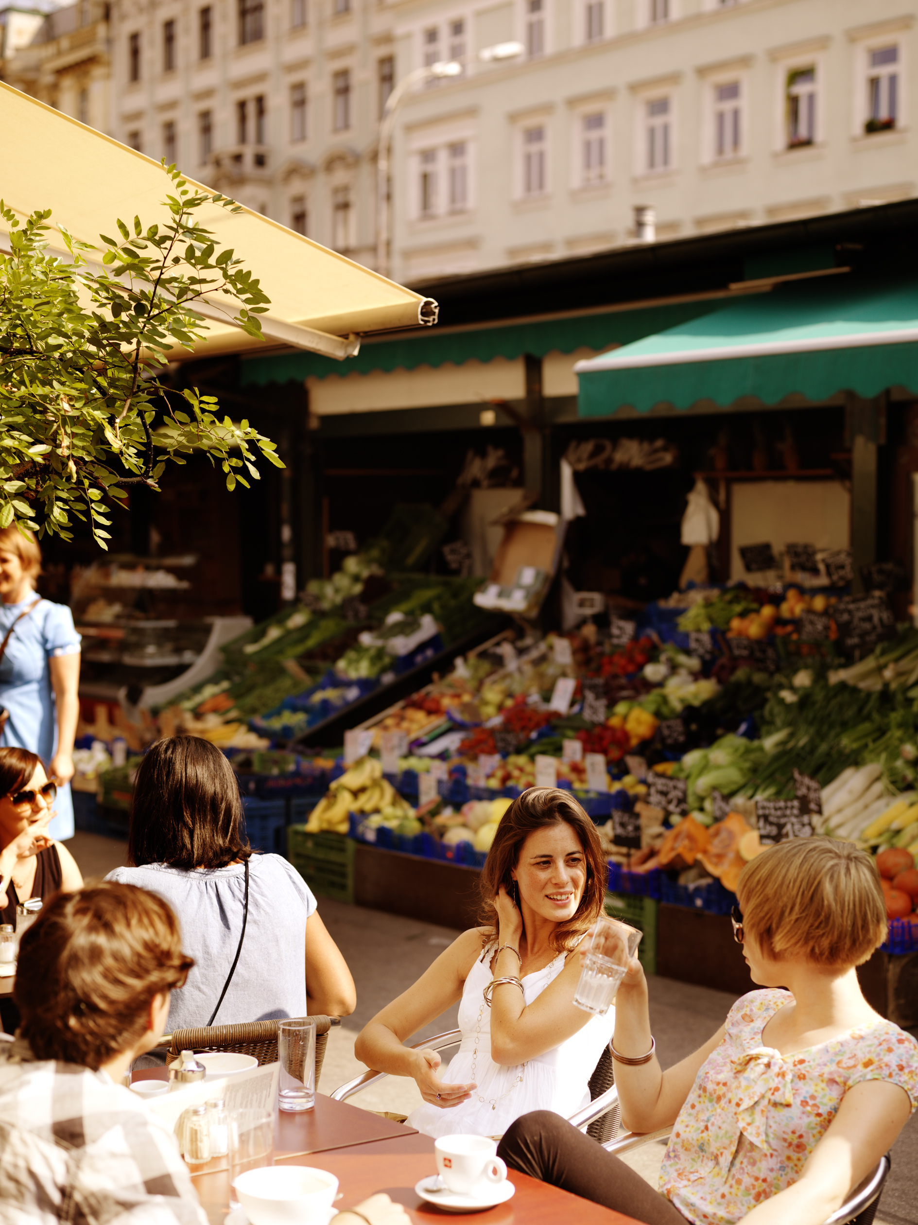 Im Vordergrund eine Gruppe Menschen die sich unterhalten während im Hintergrund das Marktgeschen mit vielen Obstständen weiterläuft.