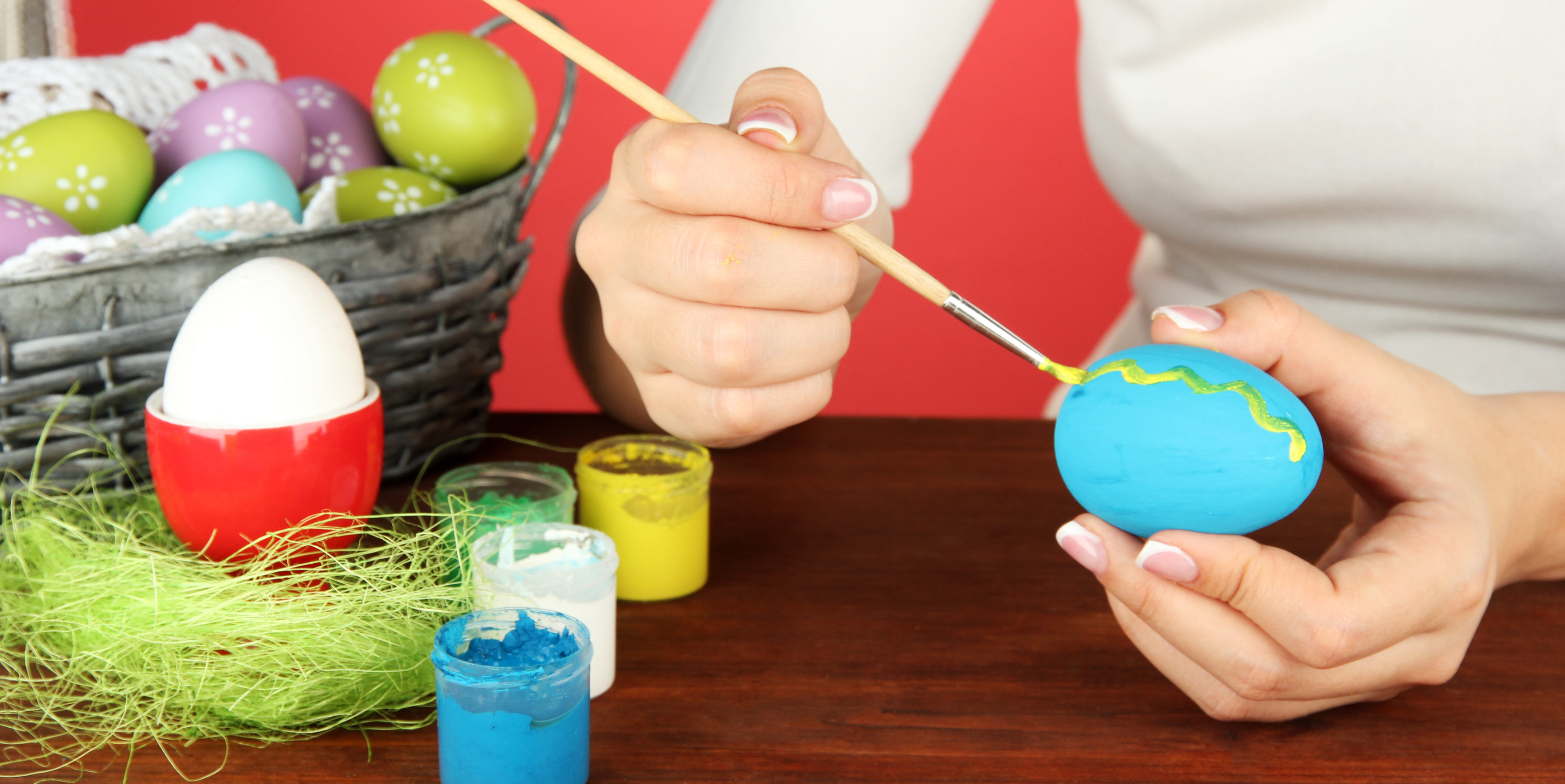 Frau bemalt mit Pinsel ein Ei blau, das sie gerade in der Hand hält. Im Hintergrund ist ein Osternest mit weiteren bunt bemalten Eiern zu sehen.