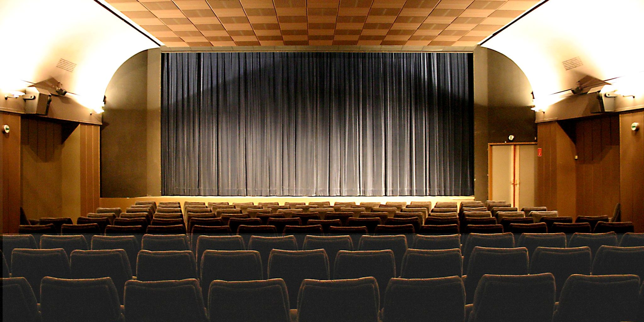 Ein braun eingerichteter Kinosaal, mit Holzdecke und einer Leinwand, die von einem grauen Vorhang verdeckt wird.