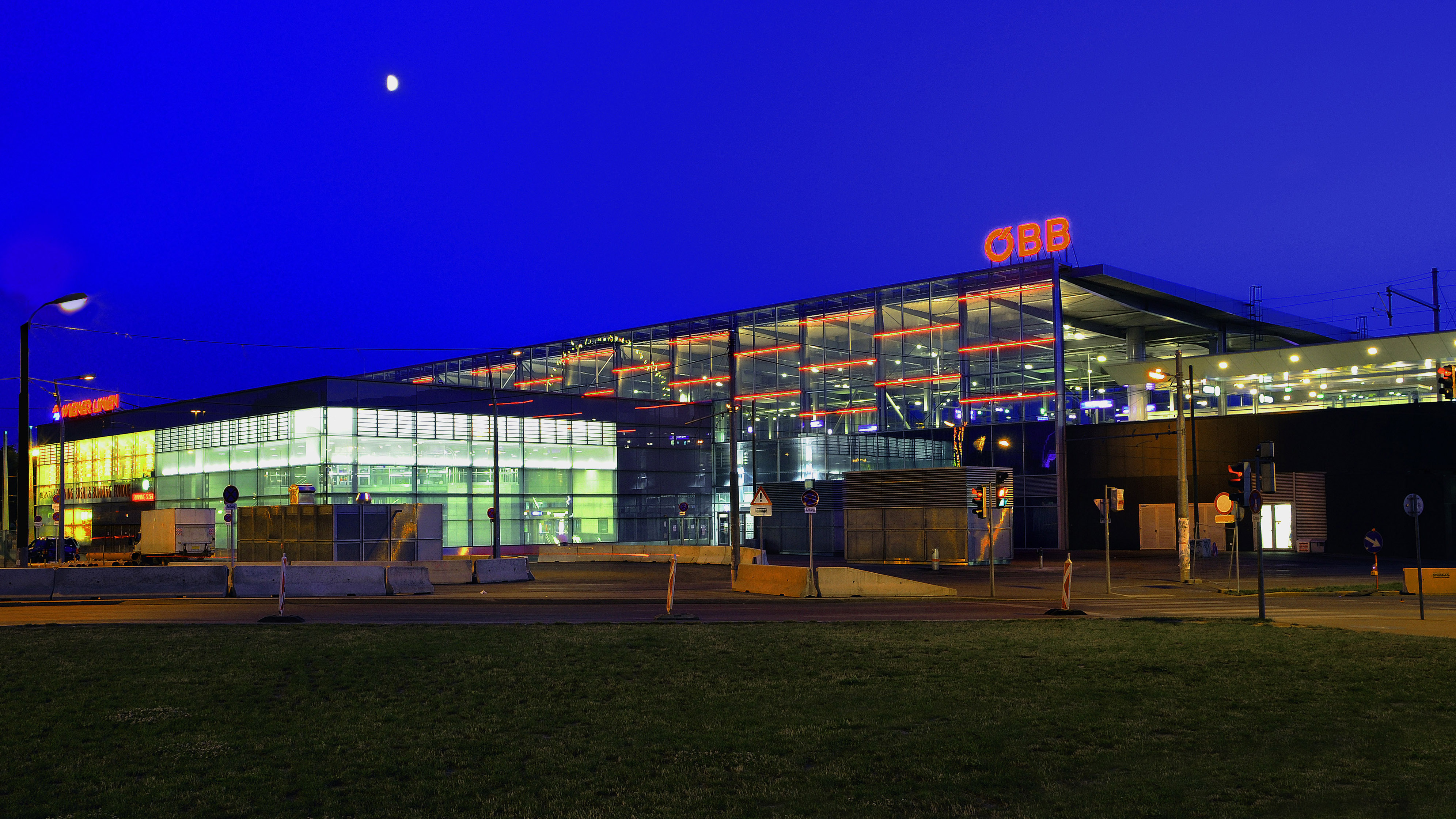 Ansicht des Bahnhofs Praterstern bei Nacht: das Gebäude ist hell erleuchtet, der Mond scheint und die Straße davor ist leer.