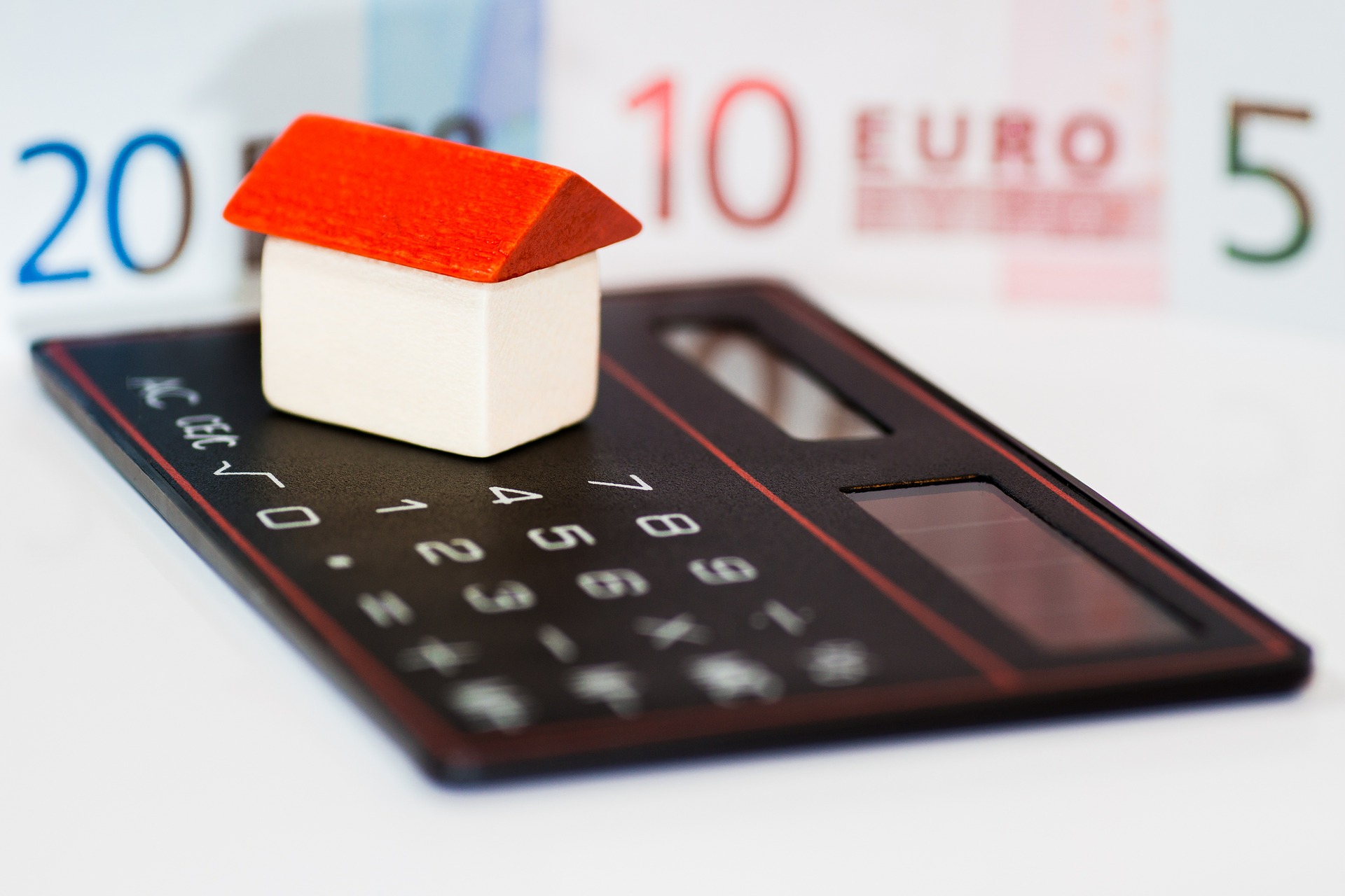 Ein kleines Hausmodell mit rotem Dach steht auf einem Taschenrechner, mit Euroscheinen im Hintergrund.