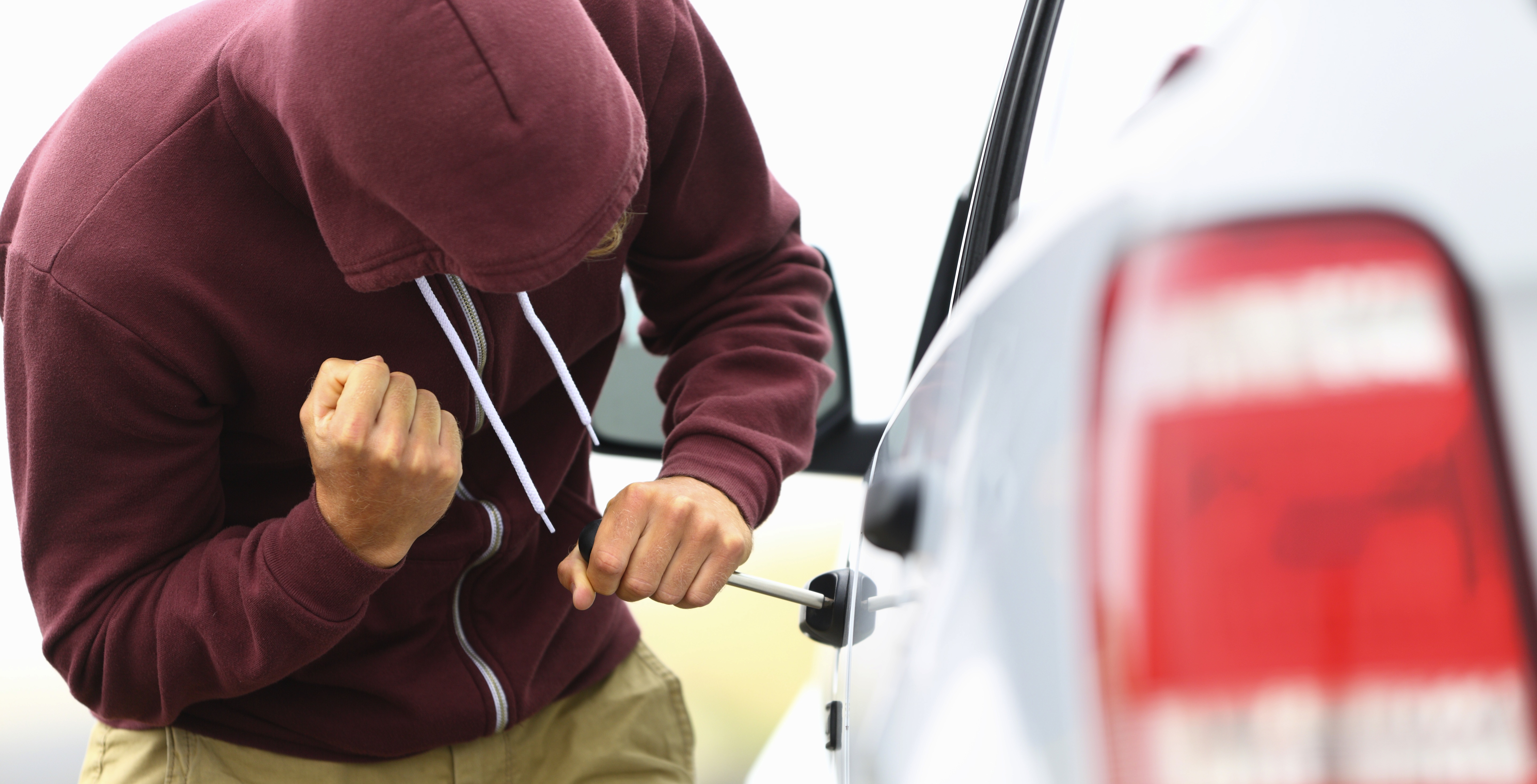 Person mit weinroter Kaputzenjacke versucht ein Autotür mit einem Schraubenzieher zu öffnen