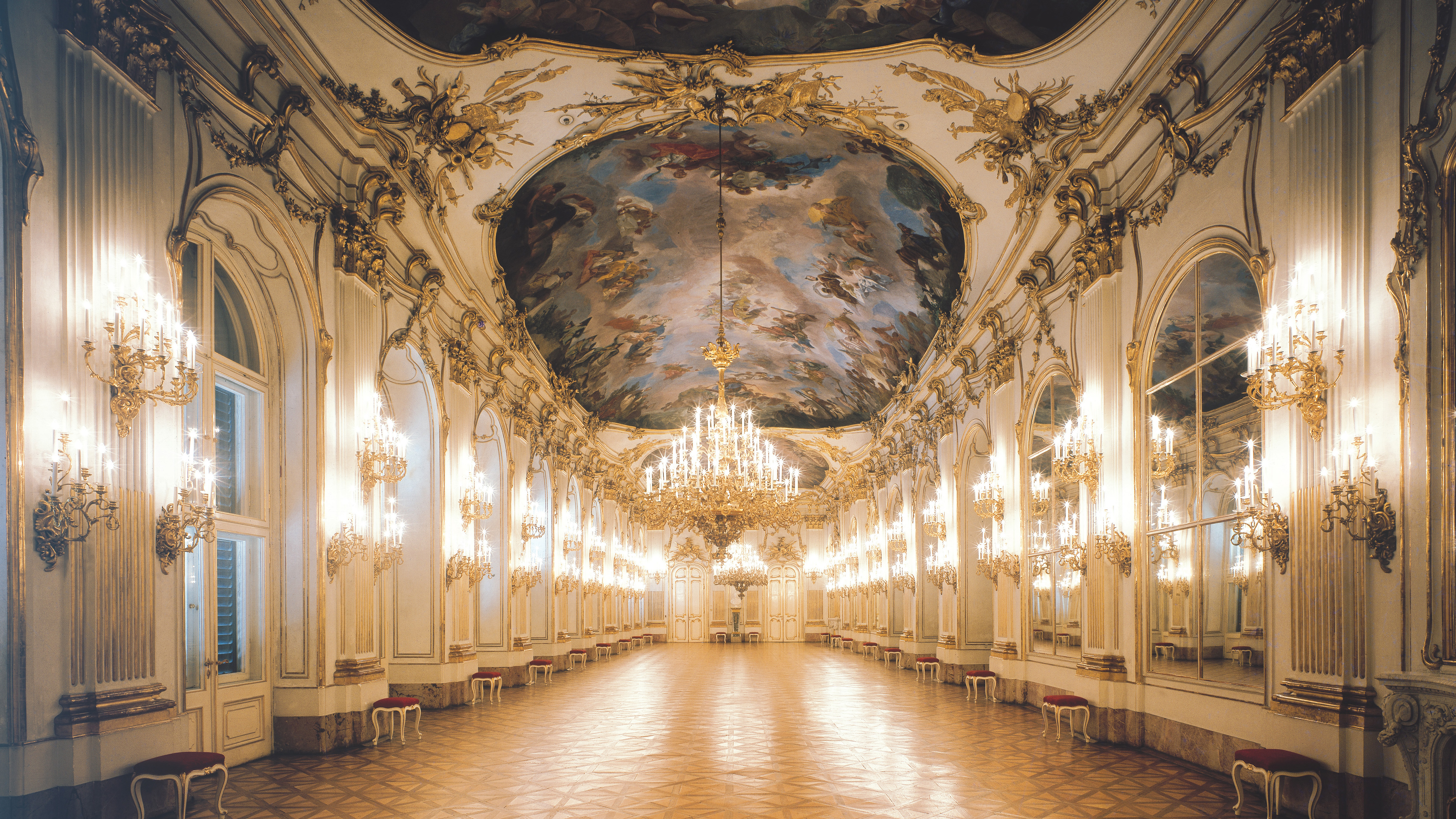 Innenansicht des Schlosses Schönbrunn: die prunkvolle Große Galerie wird von einem großen Kronleuchter in der Mitte und von vielen kleinen am Gang hell erleuchtet. 