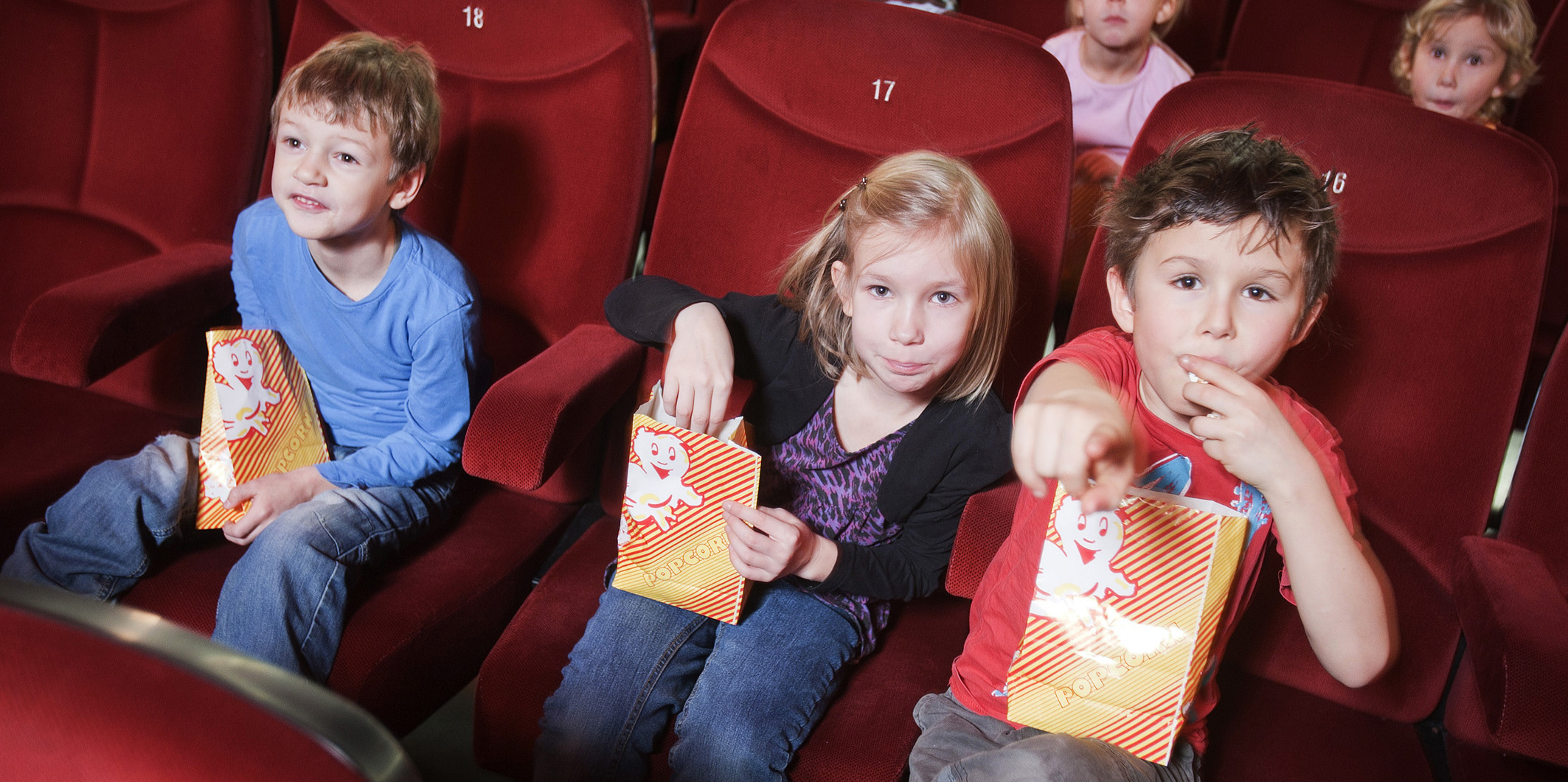 Drei Kinder sitzen in roten Kinositzen und essen dabei Popcorn.