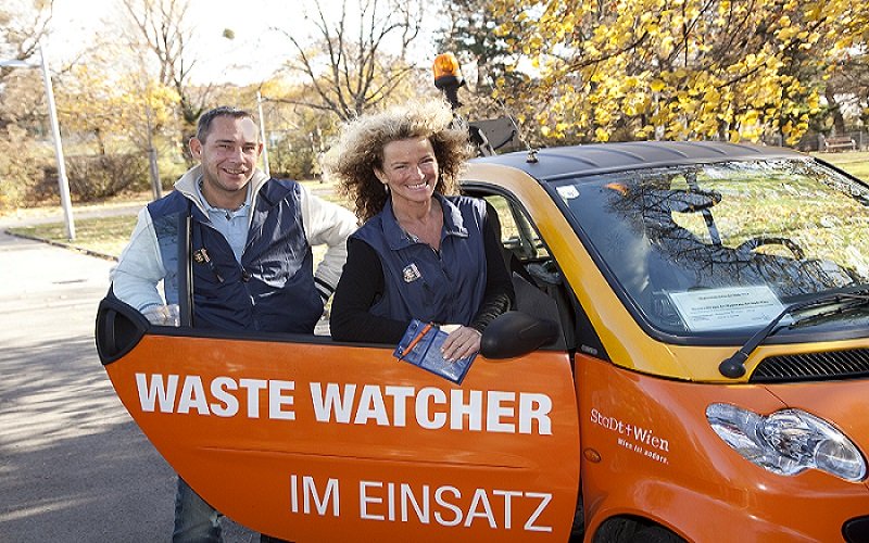 Zwei Personen, die für Waste Watcher arbeiten steigen in ein oranges Auto ein, auf dem "Waste Watcher im Einsatz steht".