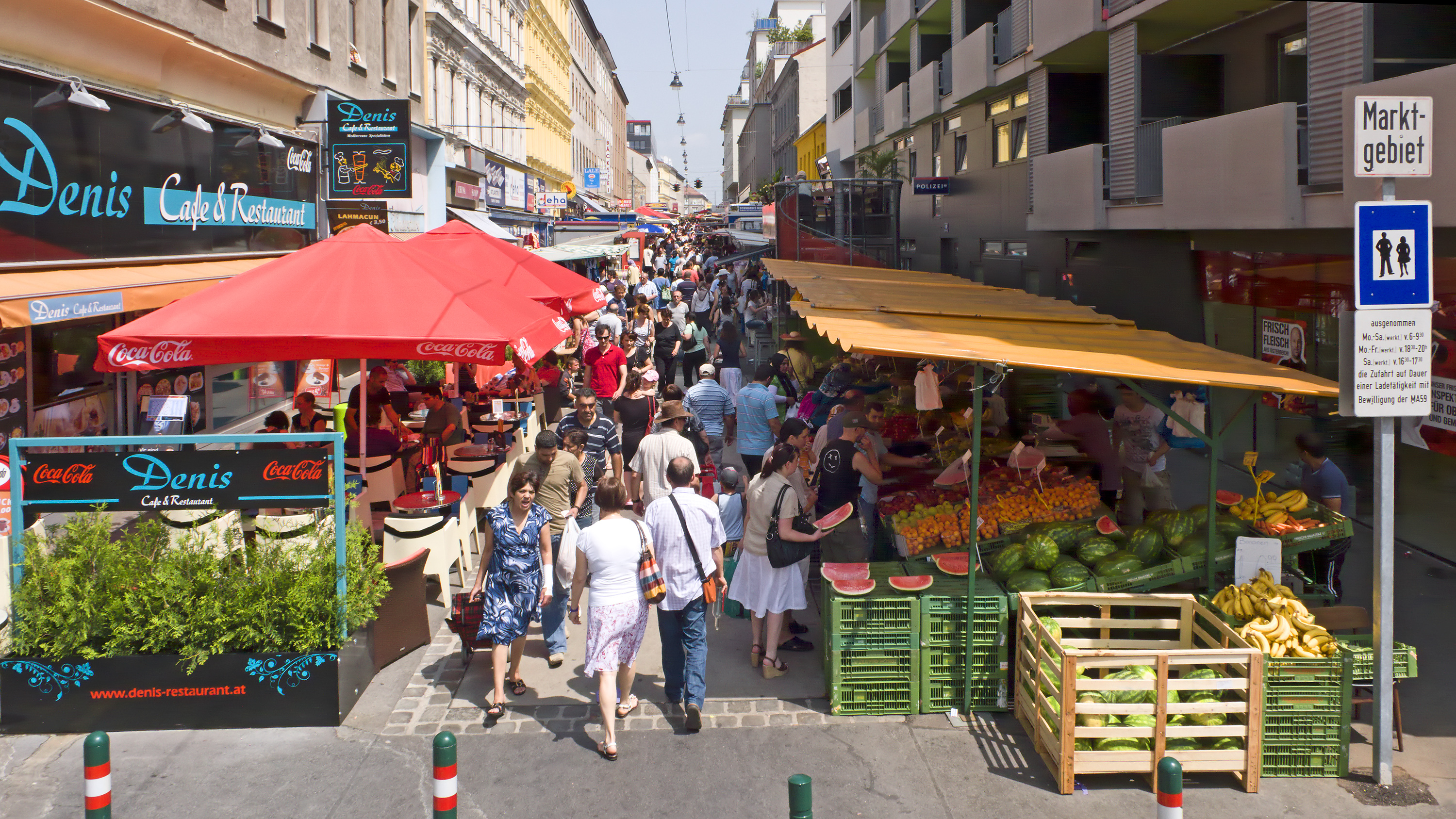 Bild vom Eingang des Brunnenmarktes: Lokale und Verkaufsstände auf beiden Seiten, letztere bieten frisches Obst und Gemüse an.