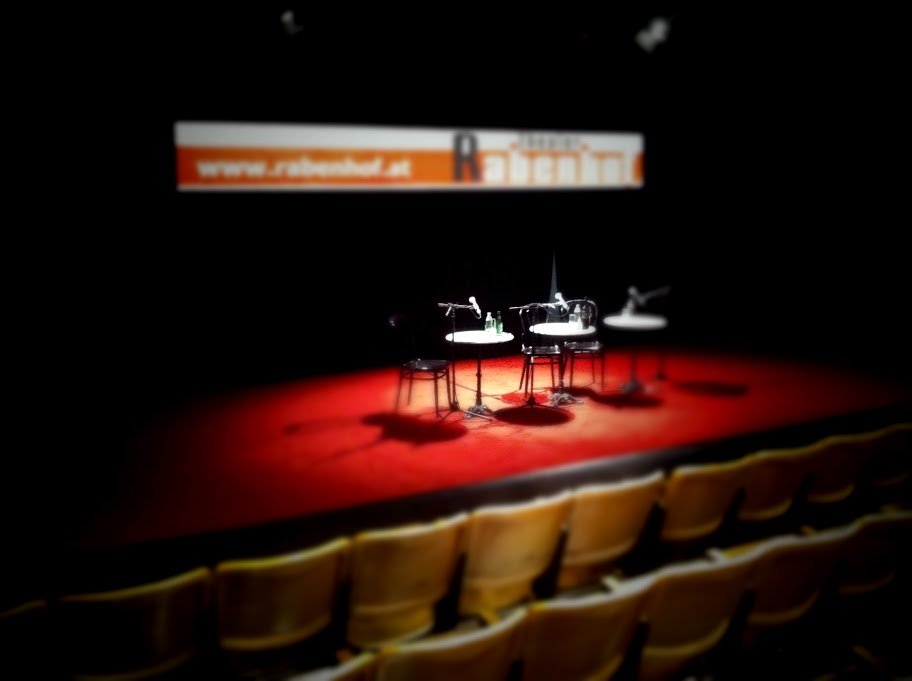 Bühne des Rabenhof Theaters Wien mit Tisch und Sesseln in rotem Licht beleuchtet
