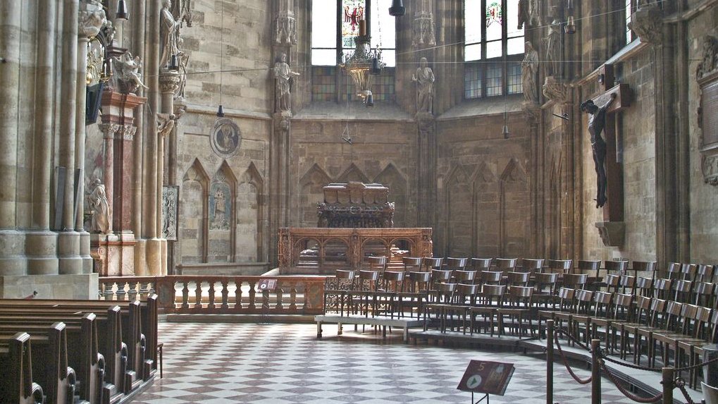 Stephansdom von innen, mit Blick auf das Grab von Friedrich III.: links stehen Kirchenbänke, rechts hängt ein Kreuz.