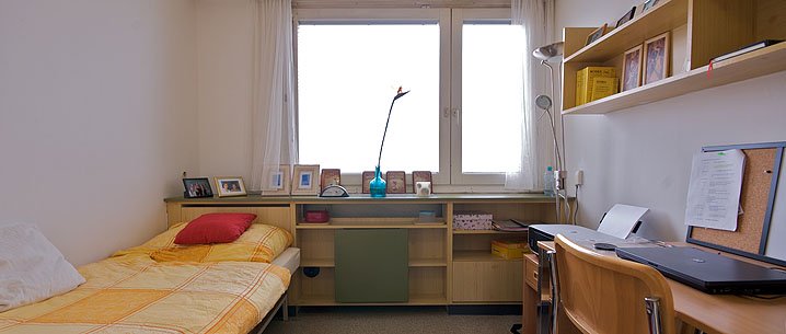 Einzelzimmer im base19 Studentenwohnheim. Einzelbett mit gelb karierter Bettwäsche und rotem Kissen. Großes Fenster. Schreibtisch, Regale und Schreibtischstuhl aus hellem Holz.