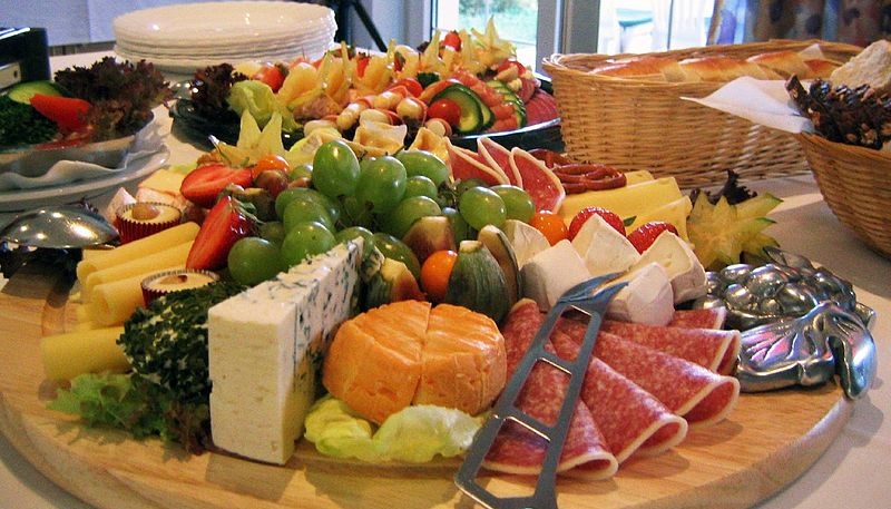 Große Platten mit Käse, Wurst, Obst und Gemüse. Dahinter Brot und Teller.