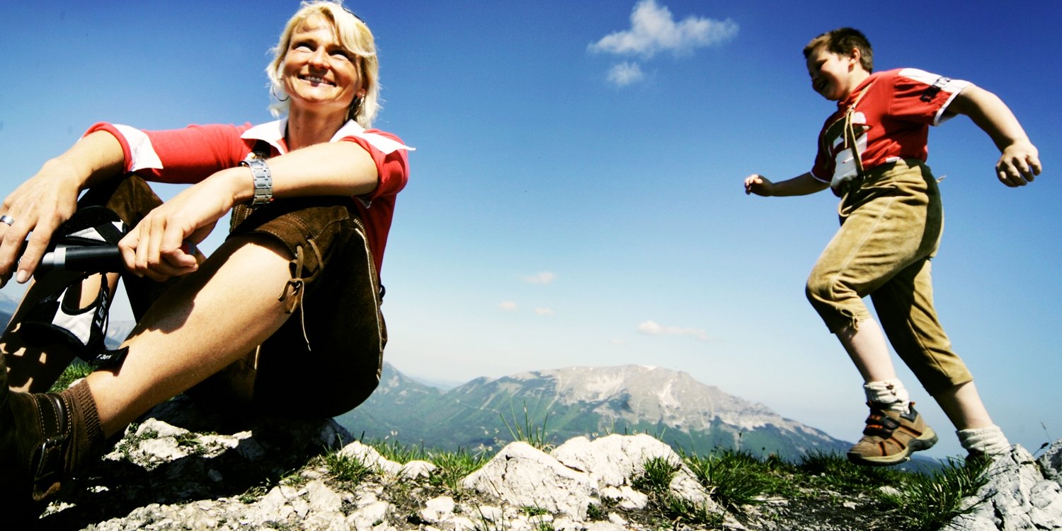 Lächelnde Frau auf Bergweise, hinter ihr läuft ein Junge in Lederhosen, dahinter Gebirgslandschaft