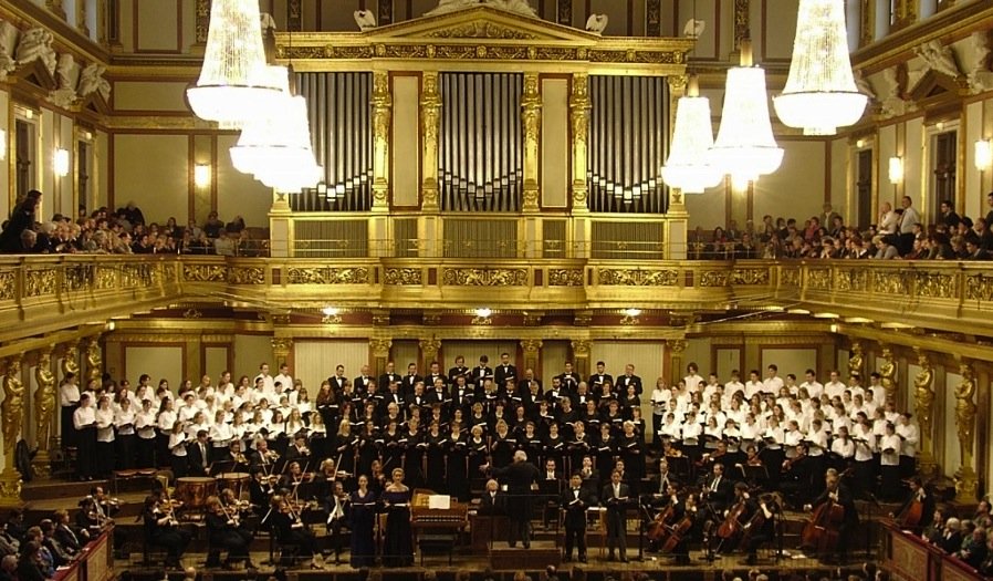 Orchesterkonzert mit Chor im Goldenen Saal im Wiener Musikverein. Publikum auf den Galerien, hell erleuchtete große Luster hängen von der Decke, im Hintergrund die goldene Orgel. 