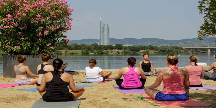 Mehrere Frauen in Yoga-Pause am Strand vom Copa Beach. Von hinten fotografiert, mit Blick auf die Donau.