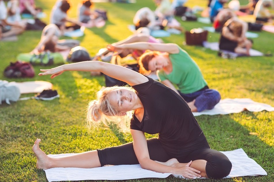 Bewegt im Park Symbolbild: Im Vordergrund Frau auf Yogamatte sitzend in Stretch-Position auf einer Wiese, im Hintergrund weitere Menschen in derselben Position