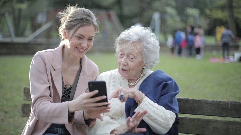 Junge Frau sitzt mit Seniorin auf Bank und zeigt ihr etwas auf dem Smartphone.
