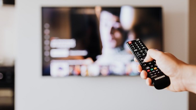 Das Bild zeigt eine Hand vor einem Fernseher, die eine Fernbedienung hält.