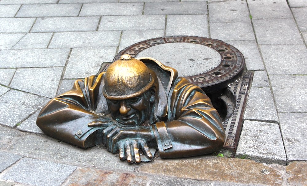 umil - Bronzeskulptur eines Mannes mit Helm, der aus einem Kanalloch schaut und den Kopf auf seinen Händen absützt